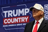 Trump, wearing a white campaign cap, poses with a stern face in front of a screen that reads Trump Country