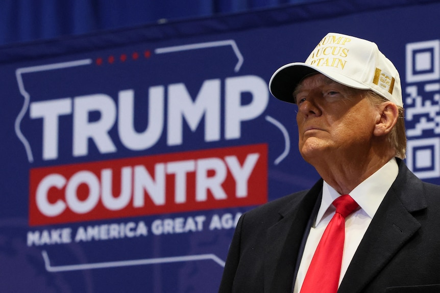 Trump, wearing a white campaign cap, poses with a stern face in front of a screen that reads Trump Country