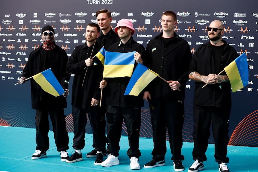A group of musicians dressed in black, holding blue and yellow flags, pose for pictures on a turquoise carpet at Eurovision.