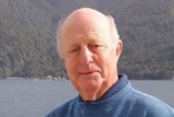 An old man leans over the railing of a boat, with a lake and hills in the background.