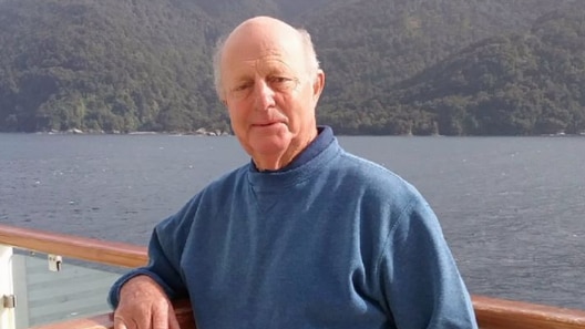 An old man leans over the railing of a boat, with a lake and hills in the background.