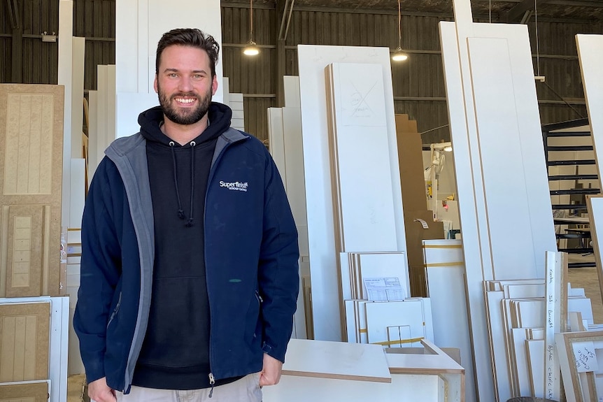 A man stands in a workshop in front of stacked kitchen joinery pieces.