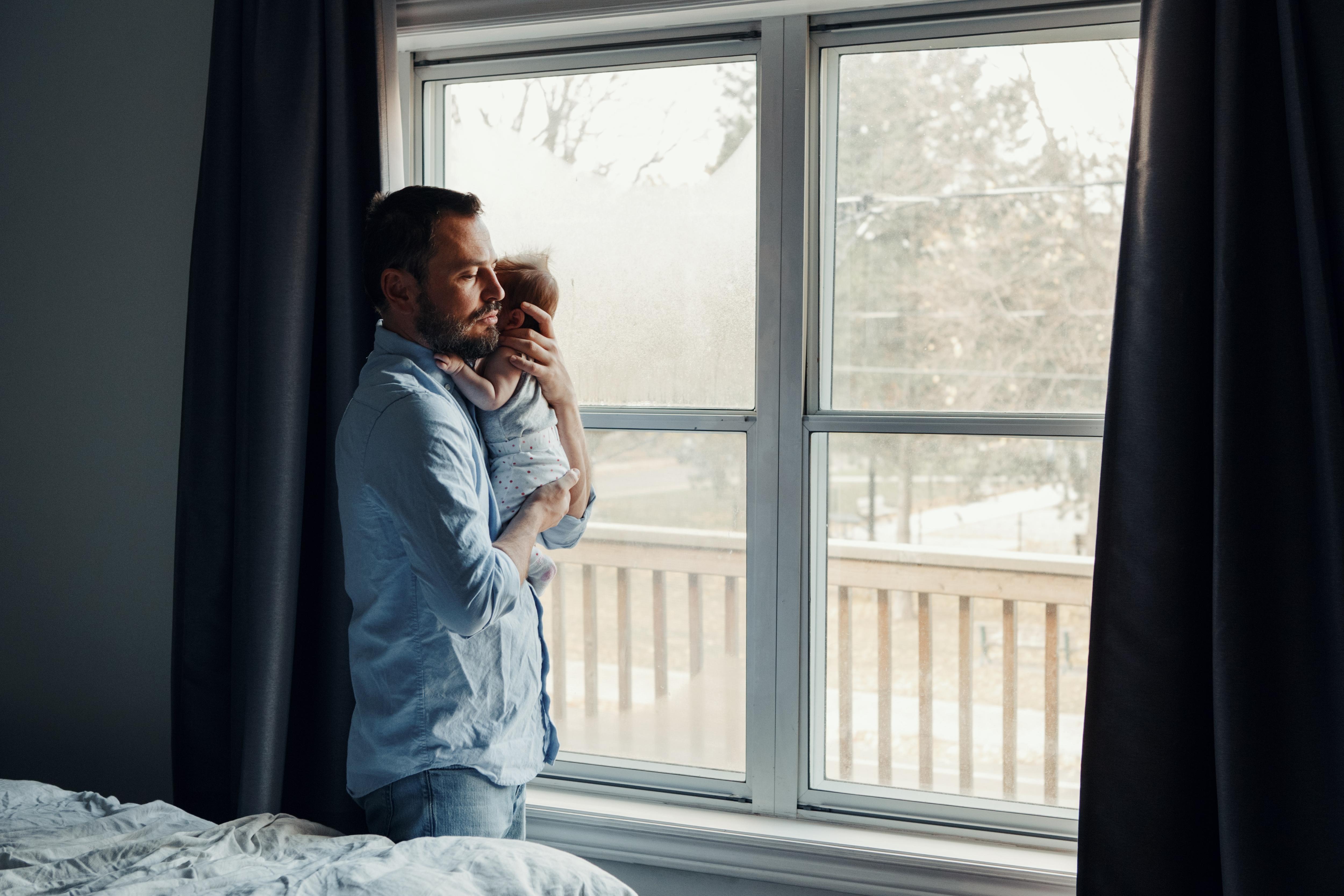 A father holds a baby, as he stands at a window.