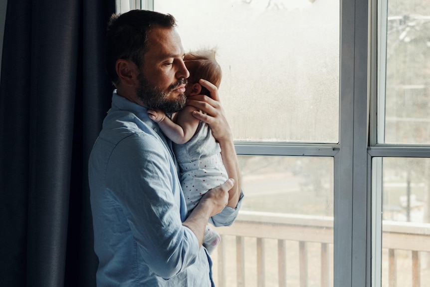A father holds a baby, as he stands at a window.