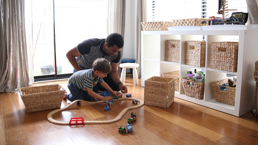 A father plays with a train set on the floor with his young son. Sun streams through the door behind them.