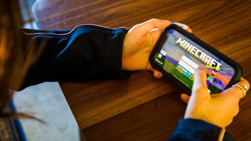 A school student holds a phone with the opening menu of the MineCraft mobile app open.