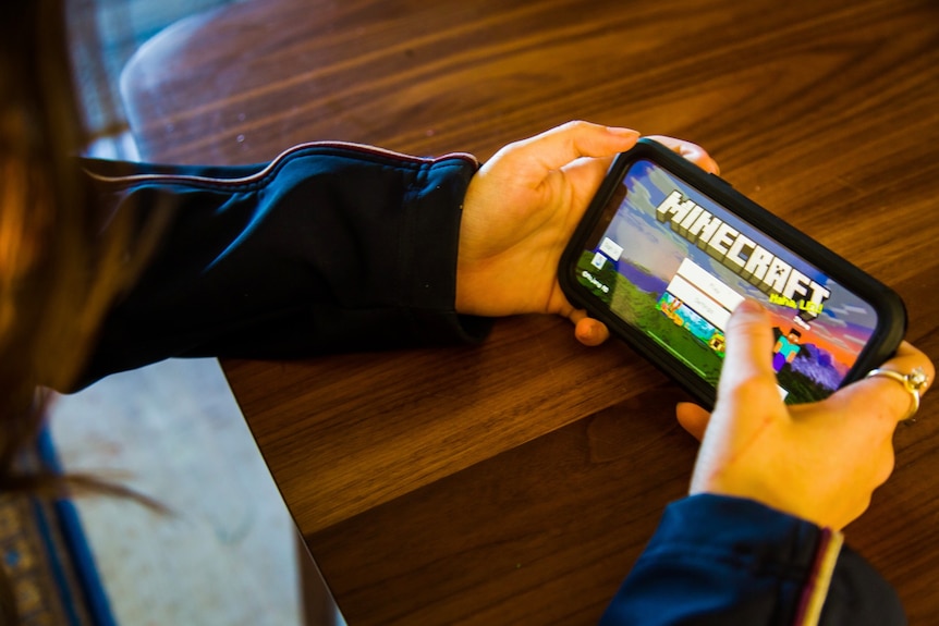 A school student holds a phone with the opening menu of the MineCraft mobile app open.