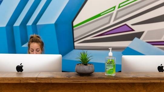 A woman sits at a desk, only part of her face can be seen over the computer screen. Hand sanitiser is on the desk.