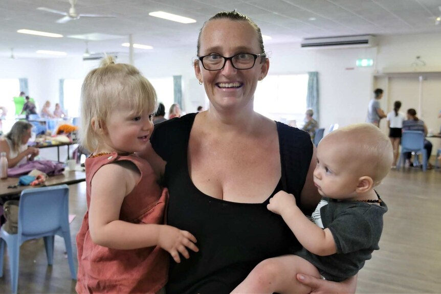Woman with glasses holding a toddler and a baby standing in a hall full of people