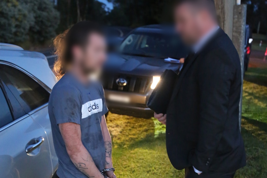 A detective speaks to a handcuffed man outside a home. Both their faces are blurred.