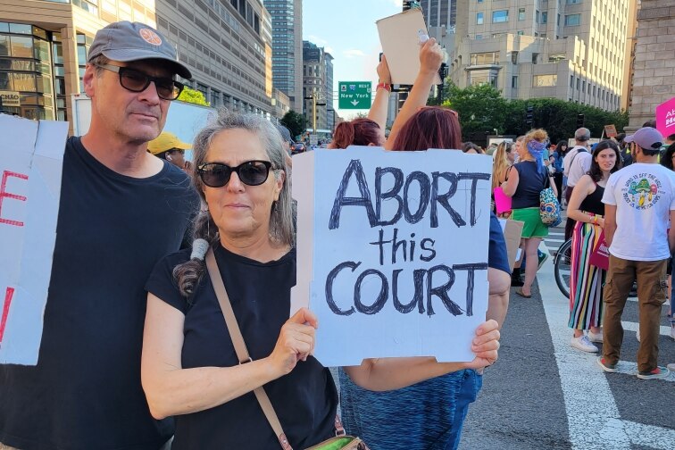 A man and a woman at a protest. The woman is holding a sign reading "Abort this Court" 