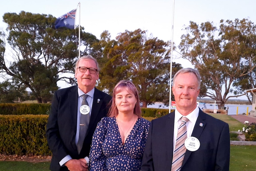 Corrina stands between the two men on a lawn by a flagpole with an Australian flag