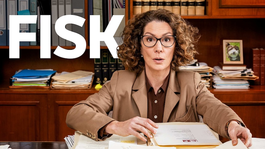 Actress Kitty Flanagan sits at a brown wooden desk with a pencil in hand, she has a curious expression