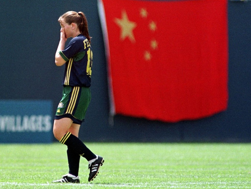 Une joueuse de football vêtue de bleu, de vert et de jaune met sa main sur son visage avec un drapeau chinois derrière elle