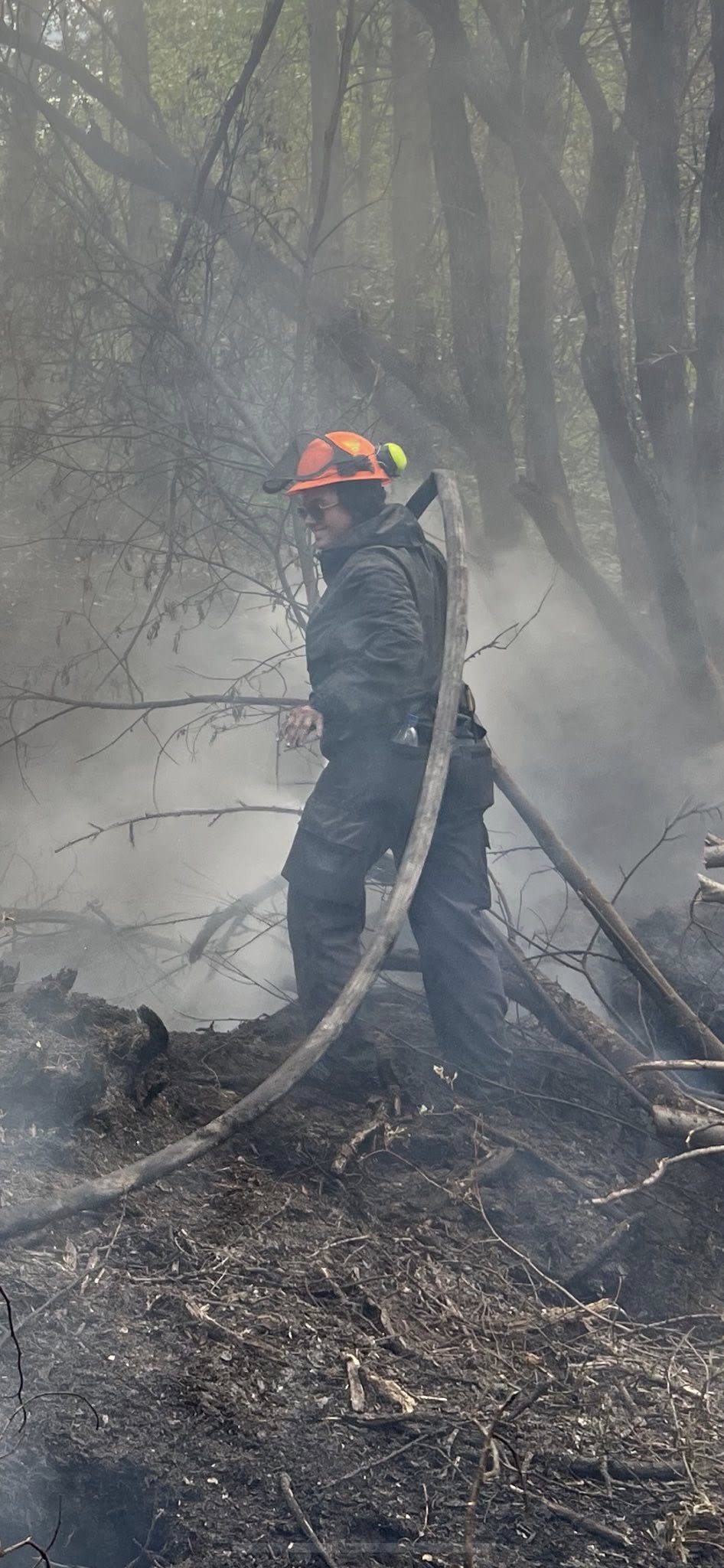 Una mujer que lleva un casco naranja y ropa negra de bombero lleva una manguera en un área llena de humo.