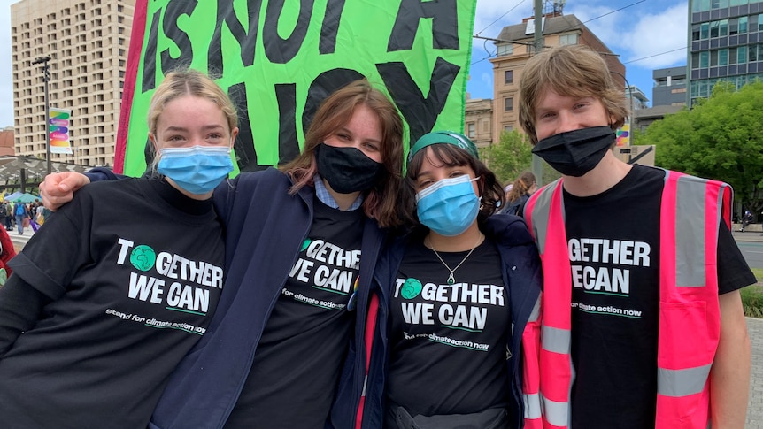 Four young people stand shoulder to shoulder wearing masks and shirts that read 'Together we can'