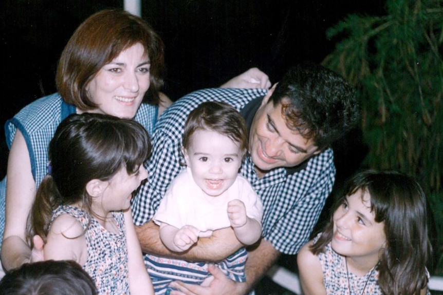 A family surround a smiling little boy in front of a table with a birthday cake on it.