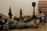 Six soldiers in fatigues lie down in front of white bust of a man, with their rifles propped up on the walls.