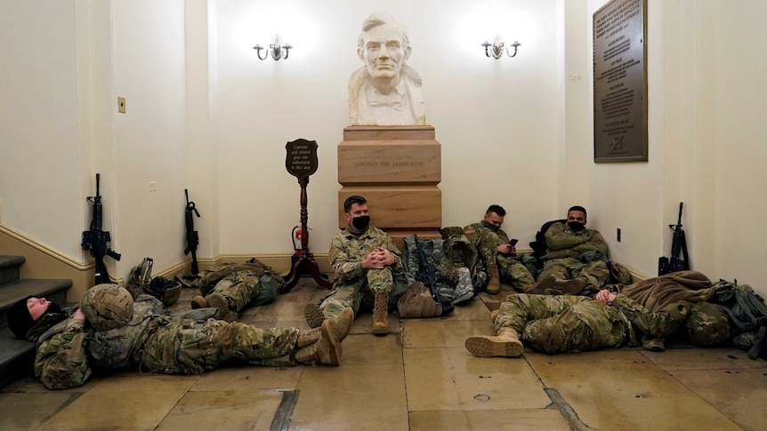 Six soldiers in fatigues lie down in front of white bust of a man, with their rifles propped up on the walls.