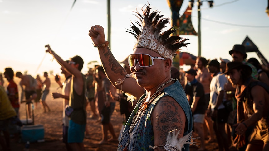 a man in a headdress and red goggles holds up his fist on the dancefloor.