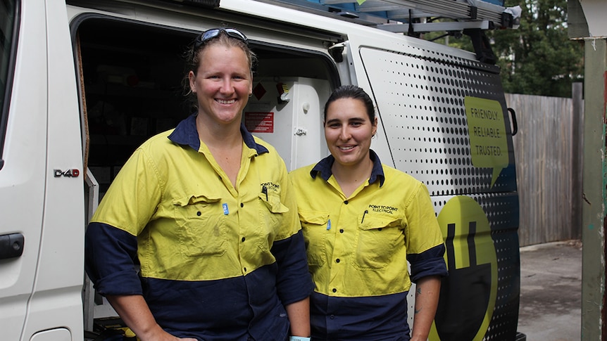 Two female electricians in blue and yellow workwear standing in front of a white van.