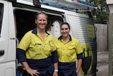 Two female electricians in blue and yellow workwear standing in front of a white van.