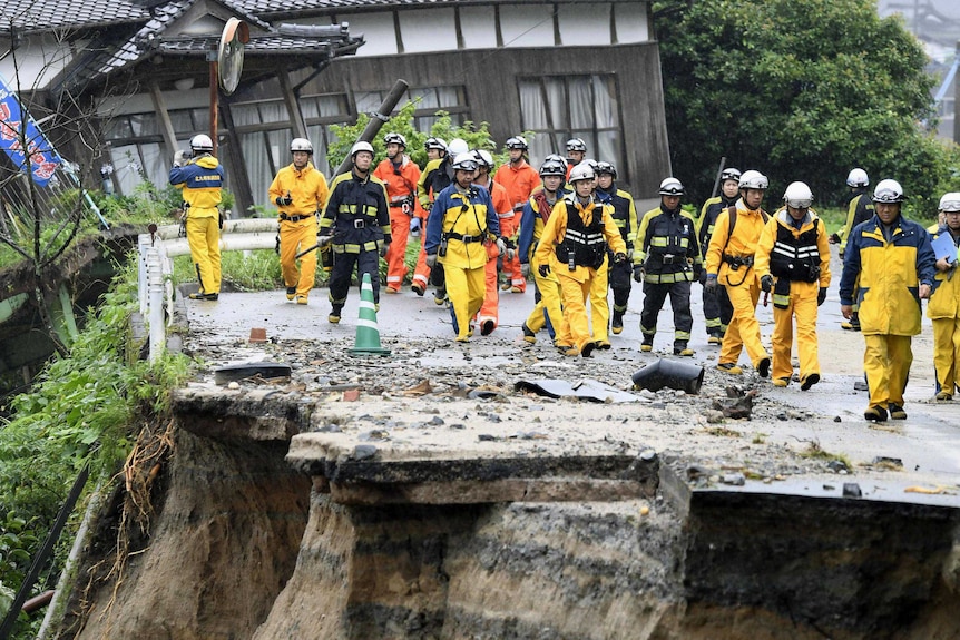 A ground of firefighters dressed in yellow walk along a damaged road which has half crumbled away because of the floods.