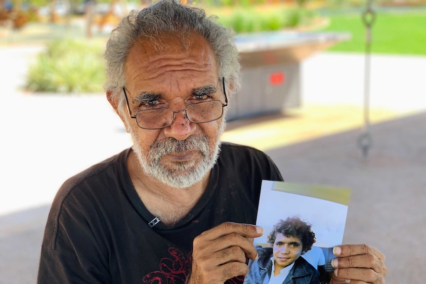 Image of a middle-aged man, wearing a black T-shirt. He's holding a hard-copy photograph of himself as a teenager..