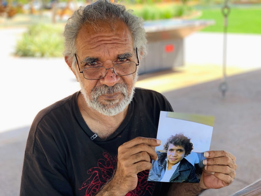 Image of a middle-aged man, wearing a black T-shirt. He's holding a hard-copy photograph of himself as a teenager..