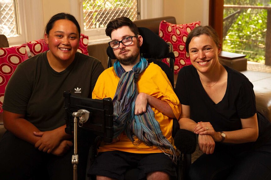 Two women pictured smiling either side of a young man in a wheelchair.