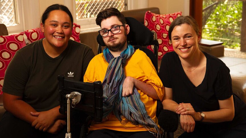 Two women pictured smiling either side of a young man in a wheelchair.