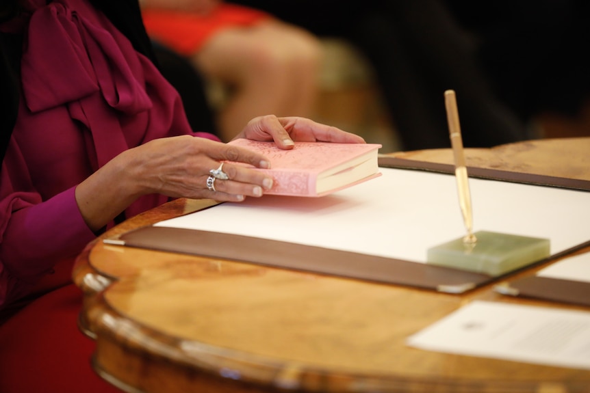 A woman holds a pink Koran with both hands as she sits at a desk with a sheet of paper and fountain pen.