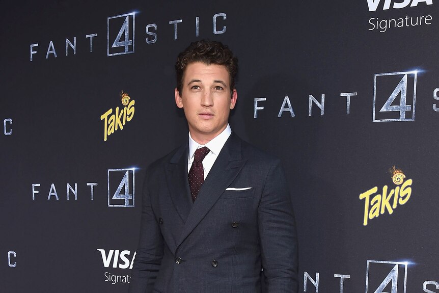 Miles Teller wearing a suit and tie, not smiling, standing in front of a wall with Fantastic 4 written on it