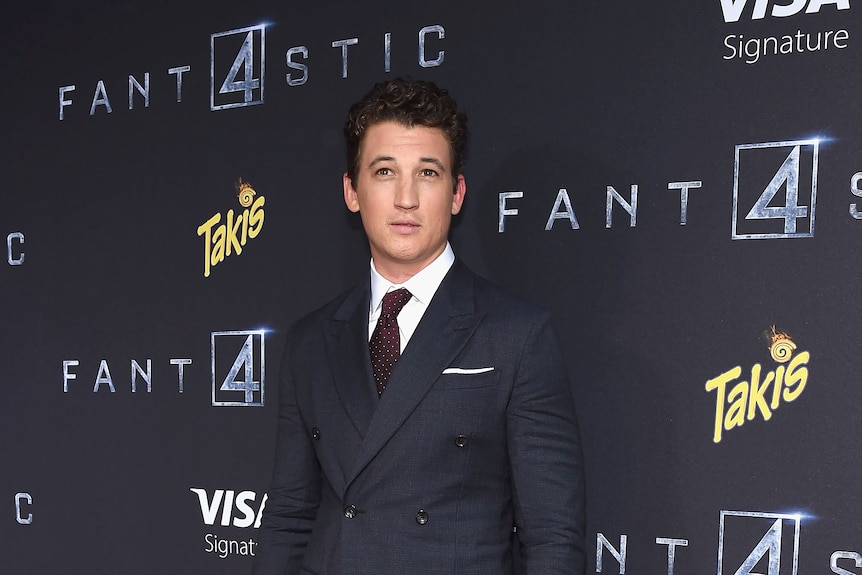 Miles Teller wearing a suit and tie, not smiling, standing in front of a wall with Fantastic 4 written on it