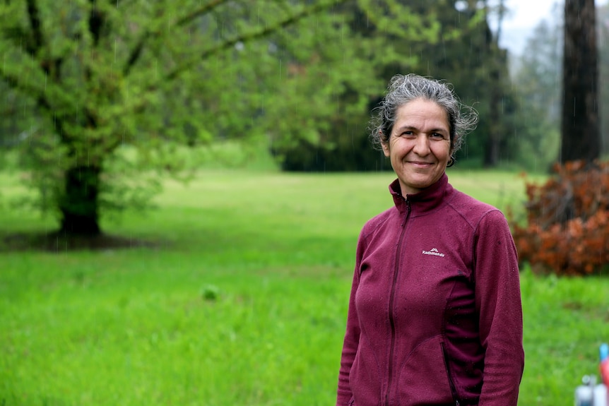 Woman in purple jumper and blue jeans with greying black hair stands outside with rain falling against tree-lined background