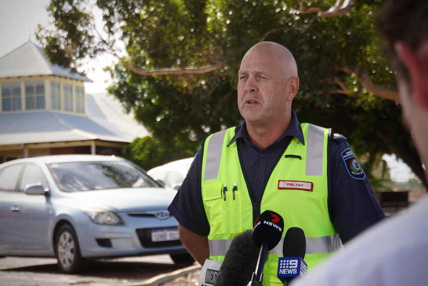 A man in a high-vis vest standing in front of microphones and talking to the media.