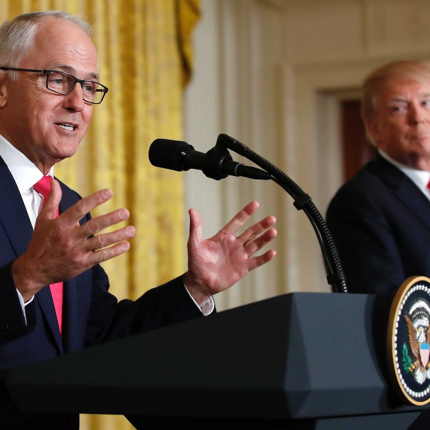 Donald Trump, in the background, gazes at Malcolm Turnbull as he speaks at a podium with his hands gesturing