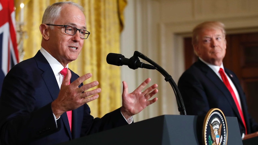 Donald Trump, in the background, gazes at Malcolm Turnbull as he speaks at a podium with his hands gesturing