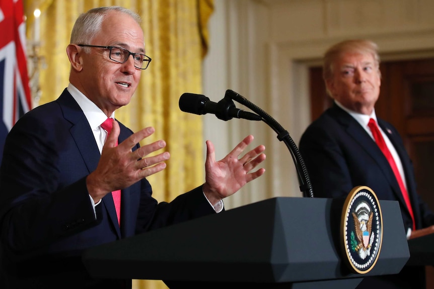 Donald Trump, in the background, gazes at Malcolm Turnbull as he speaks at a podium with his hands gesturing