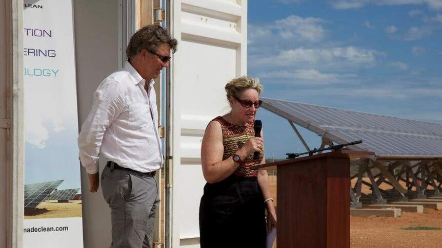 A man and woman speak at a lectern in an outback setting in front of a solar power farm.