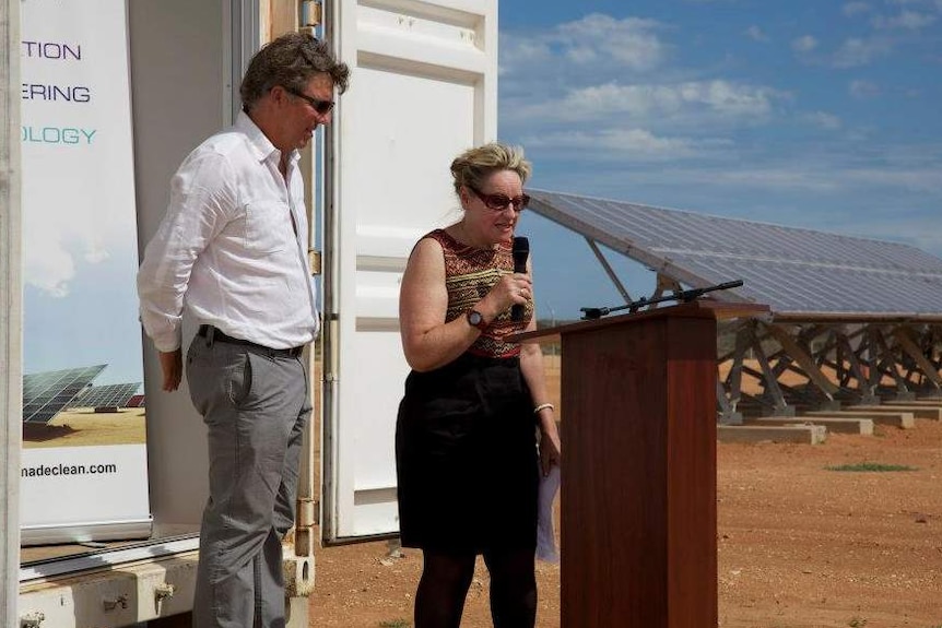 A man and woman speak at a lectern in an outback setting in front of a solar power farm.