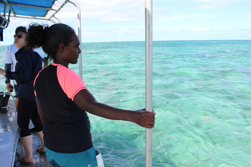 Dr Katy Soapi holding onto a pole at the edge of a dive boat looking into crystal clear water with others behind her