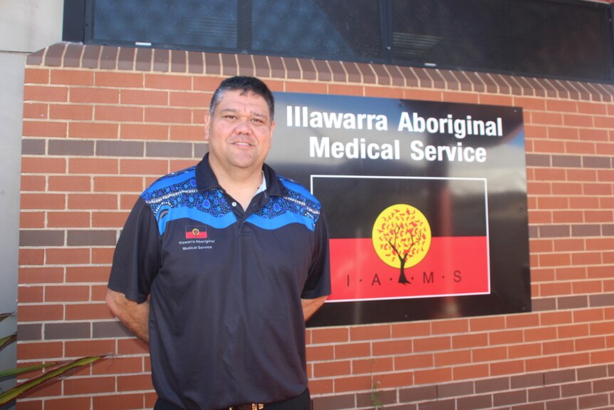 A man standing next to a sign that says "Illawarra Aboriginal Medical Service".