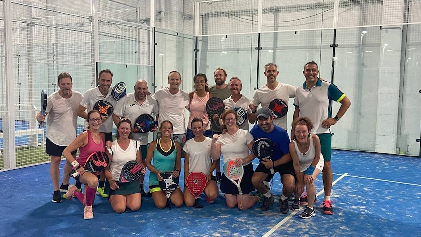 Team of people wearing white shirts in front of a Padel court holding Padel racquets.