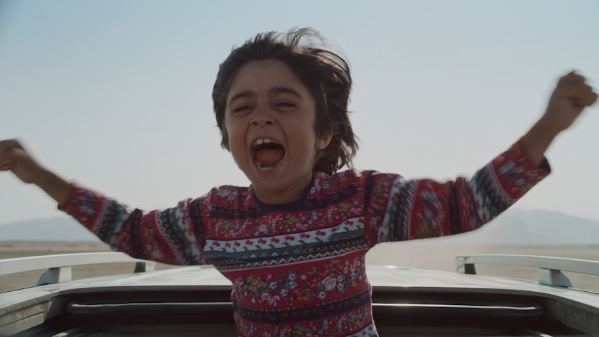 A young boy wearing a patterned red, white and black sweater is standing through a car sun roof, arms spread and yelling in joy.