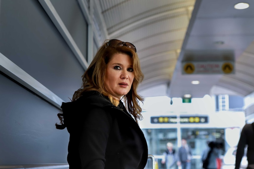 Woman in black hoodie and brown hair stares with entry to airport in background