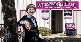 Older woman wearing a suit and with a slight smile sitting in front of a pink 'Questa Casa' sign.