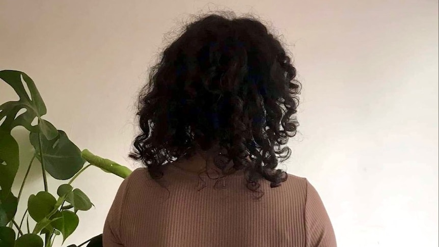 A young lady with a brown sweater and black curly hair facing the wall.