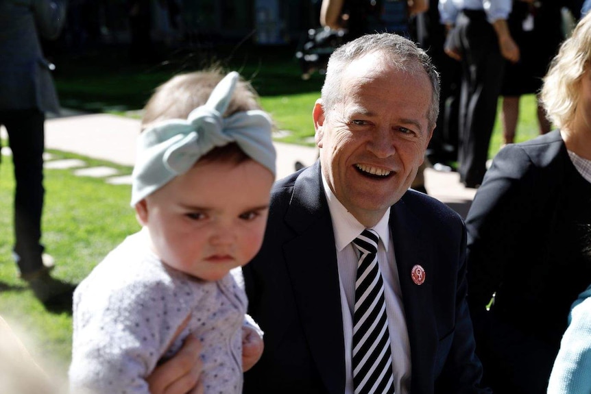 Baby Lola sits in the foreground wearing a blue head scarfe while Bill Shorten smiles at her in the background