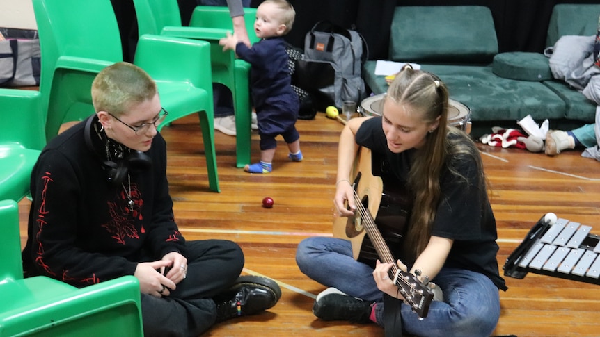 two young women sit on the floor, one has a guitar. a standing baby is in the background, holding on to a chair.